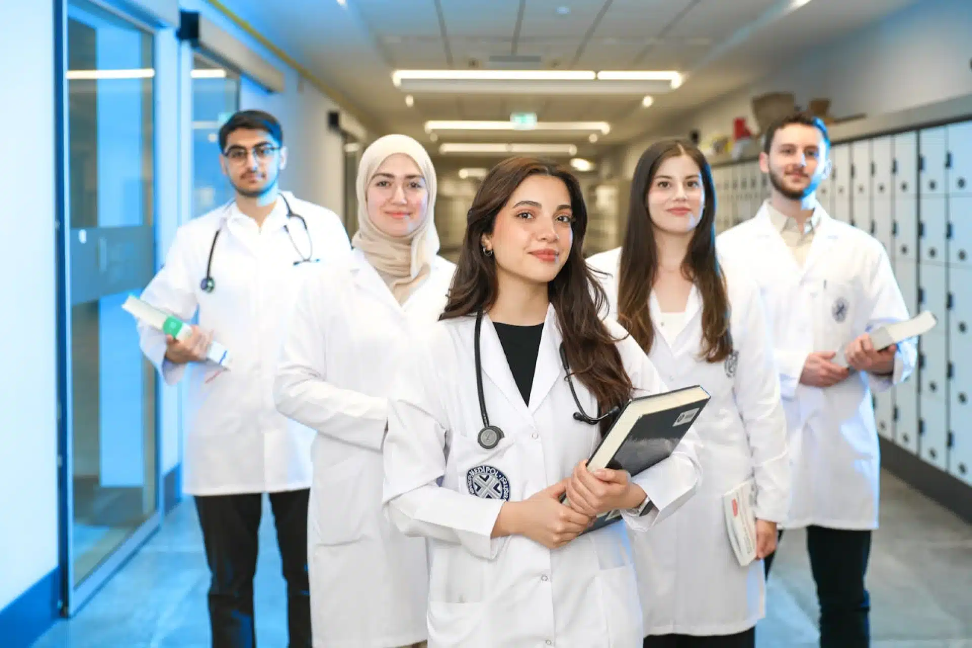 Diverse group of five healthcare students in white lab coats and stethoscopes in a hallway.