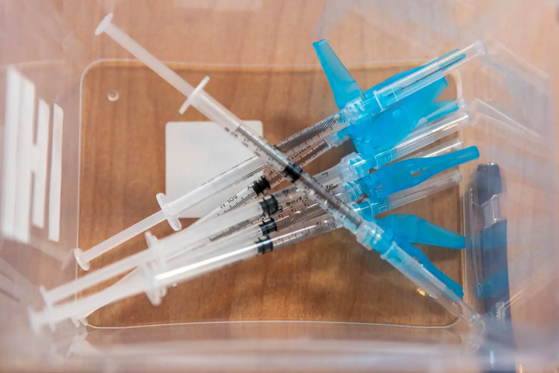 Close-up of several new, unused syringes and capped needles inside a clear plastic container.