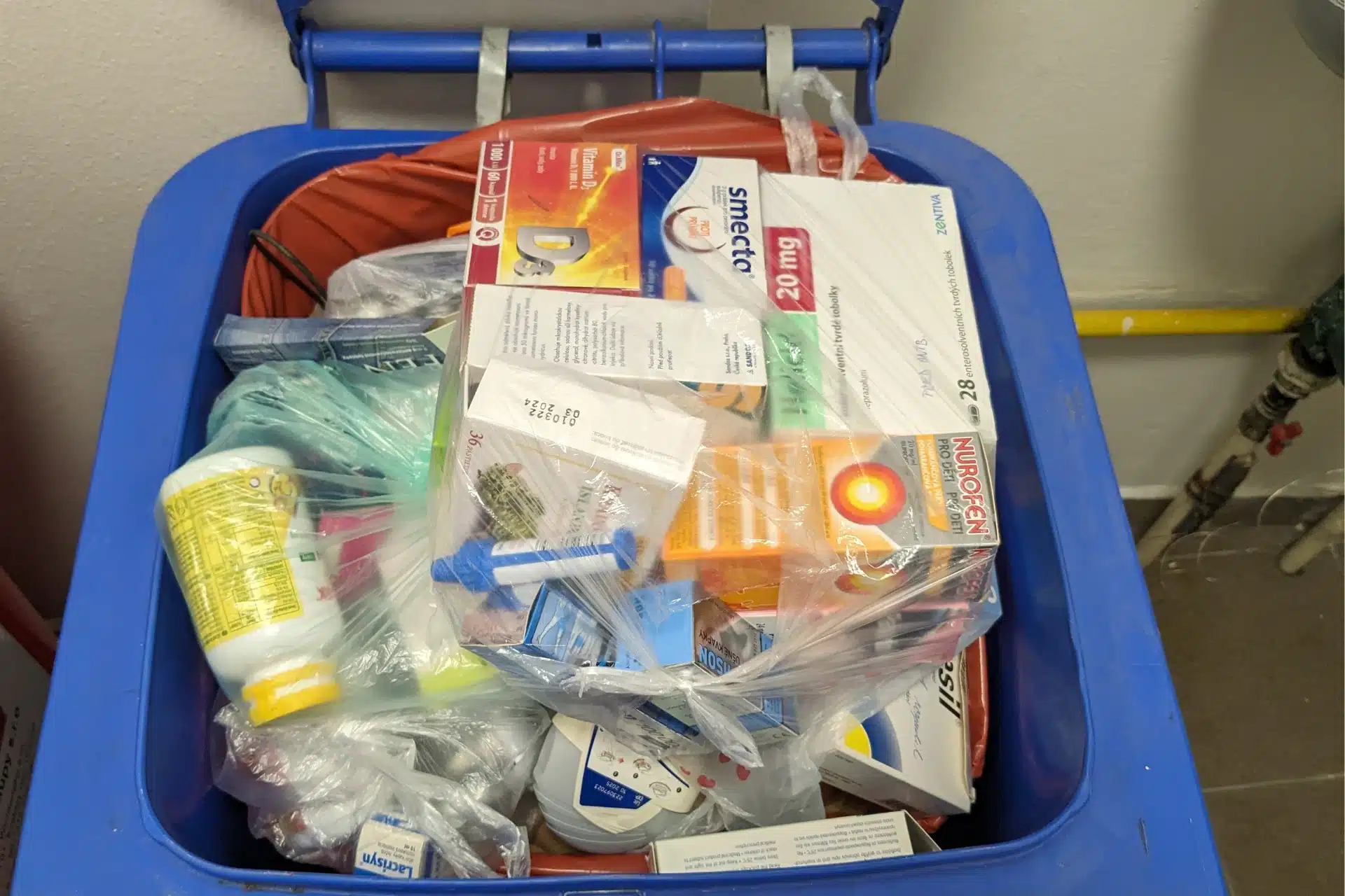 A blue disposal bin filled with a clear plastic bag containing mixed pharmaceutical waste and discarded medicine boxes 
