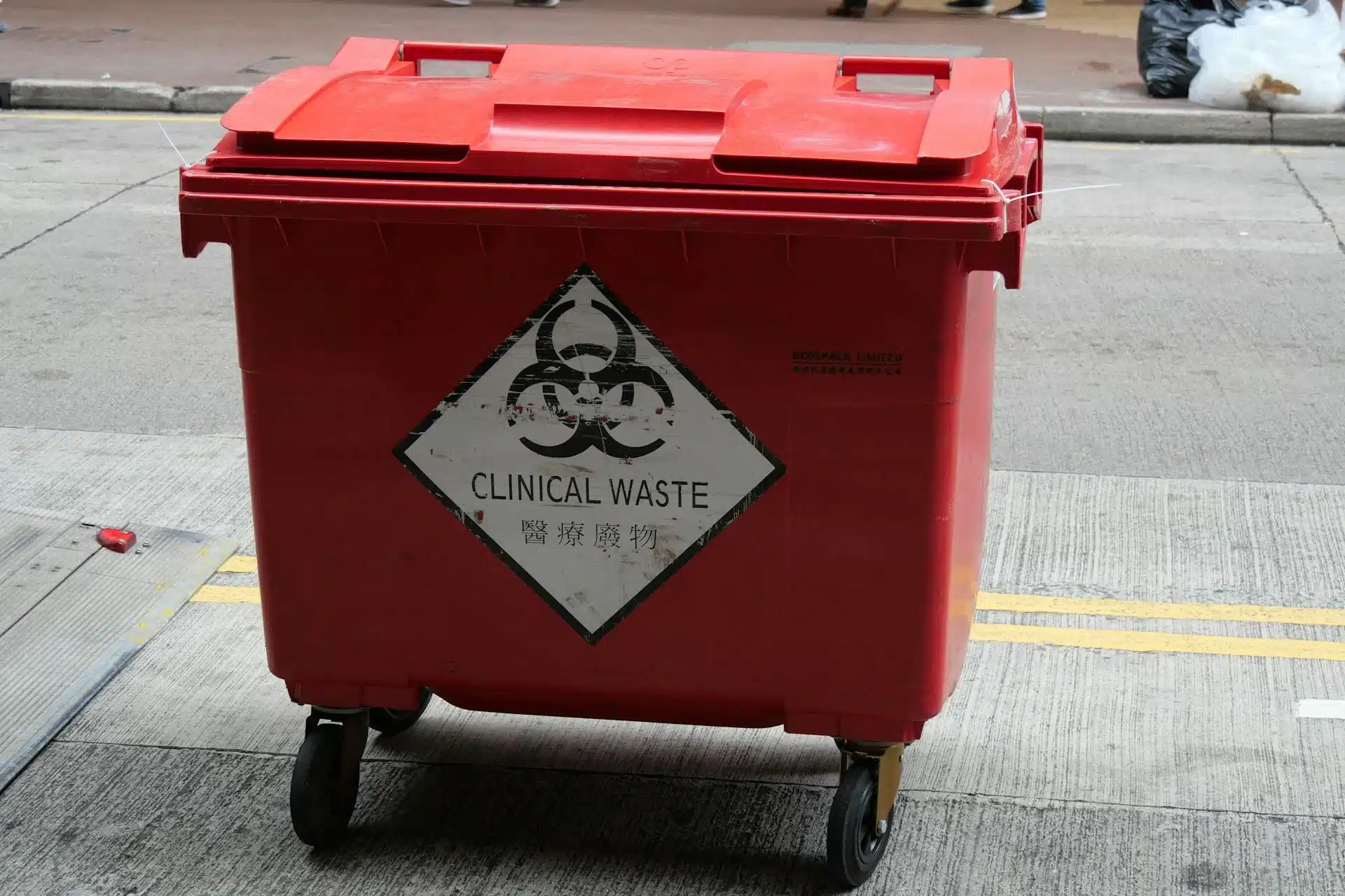 A red wheeled medical waste bin on a city street, labeled with a biohazard symbol and the words “Clinical Waste.”