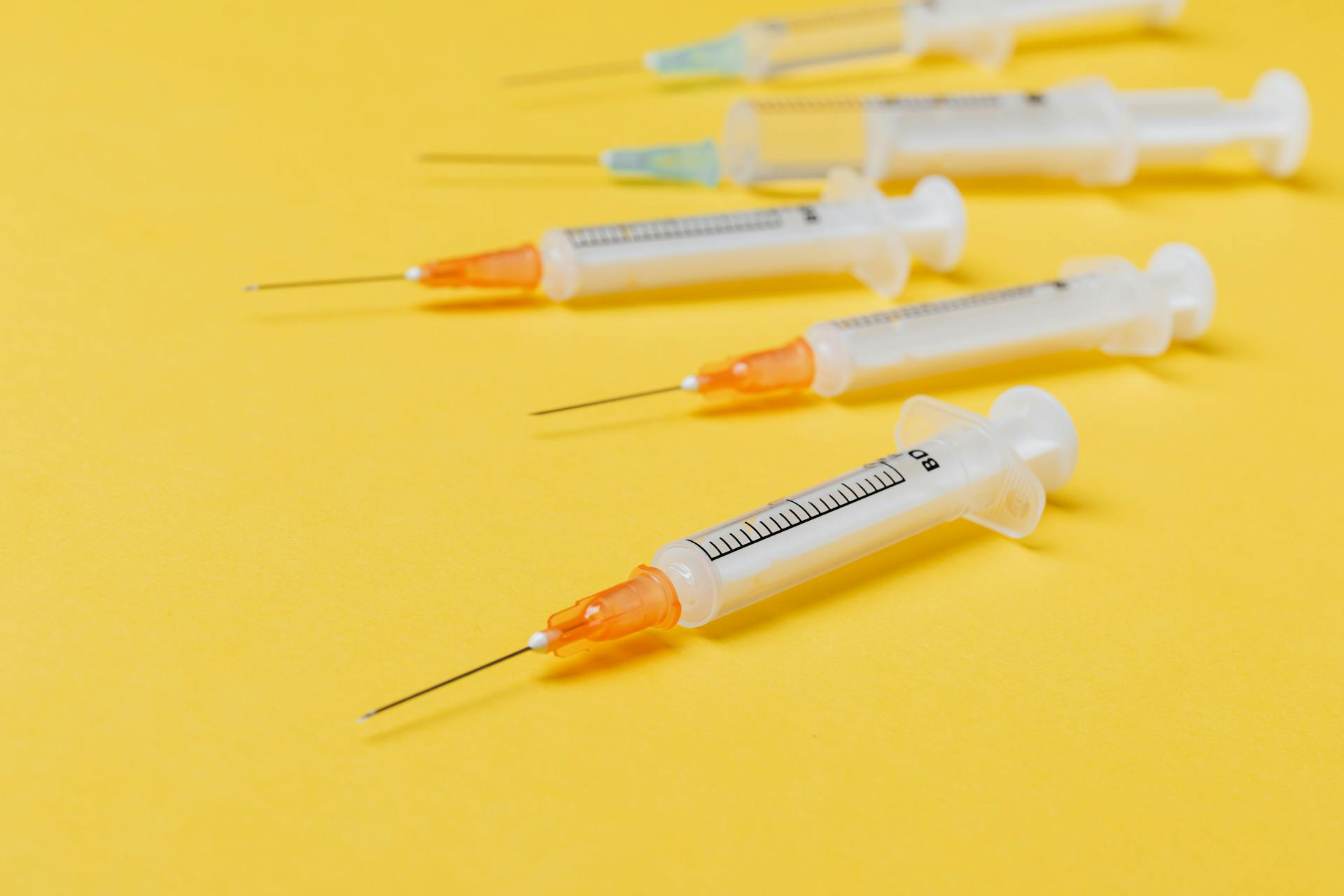 Several disposable medical syringes with needles arranged diagonally on a bright yellow background 