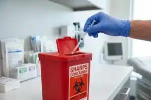 A gloved hand disposing of a medical syringe into a red FDA-cleared sharps container