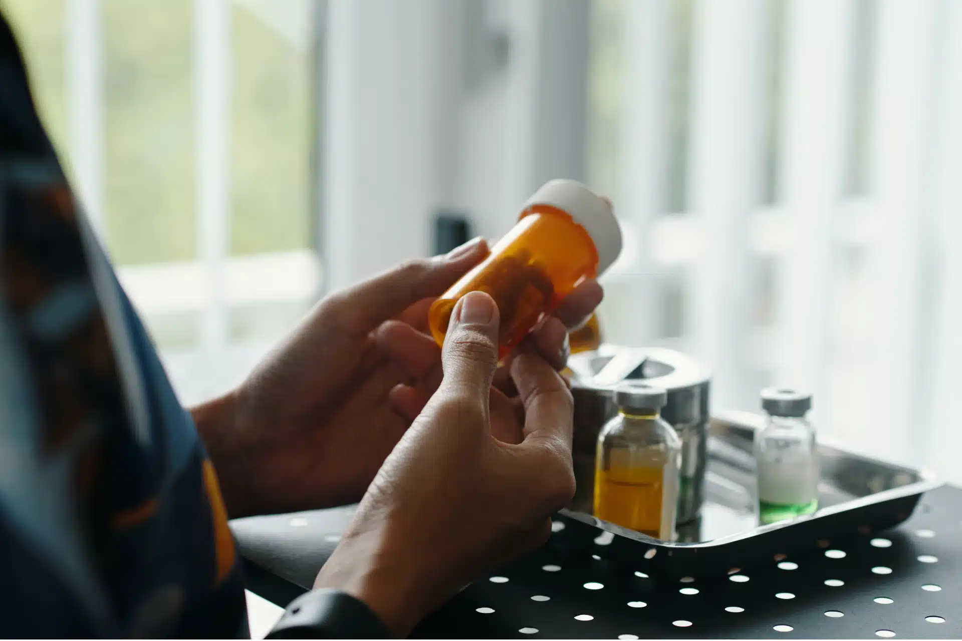 Close-up of a person holding an amber pill bottle, with small medical vials sitting on a metal tray in the background.