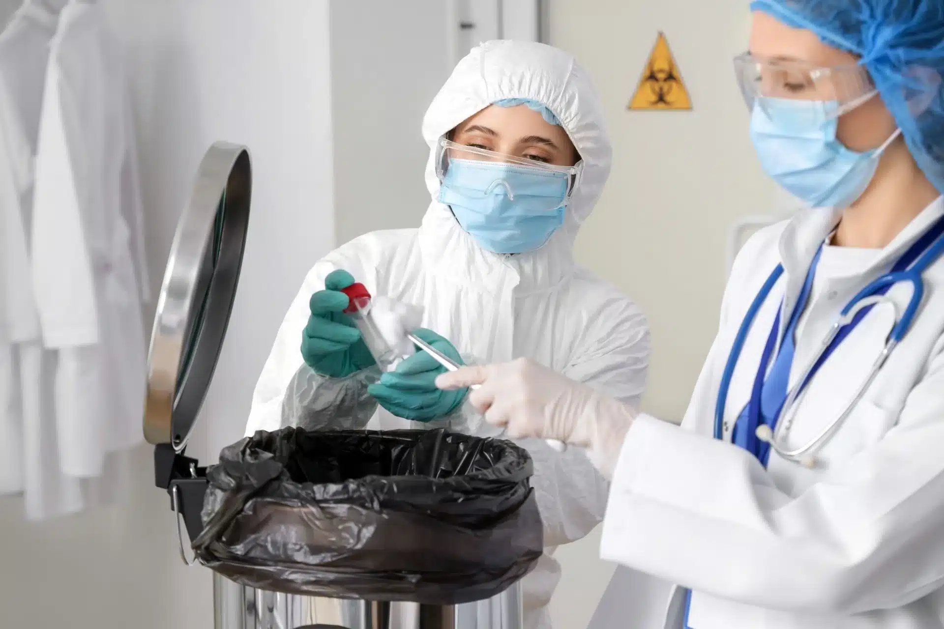 Two lab workers in protective gear handling a small container near a biohazard waste bin. 