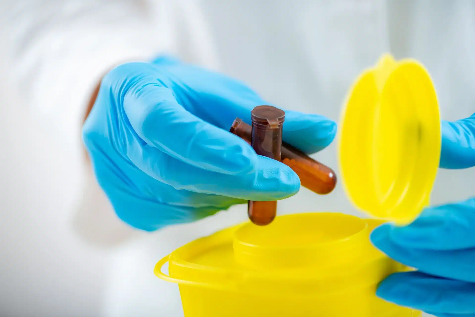 Gloved hands placing small glass vials into a yellow biohazard disposal container in a laboratory setting.